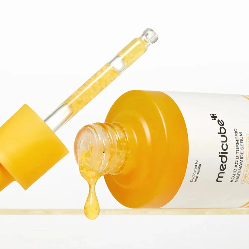 A close-up view of an open Medicube Kojic Acid Turmeric Niacinamide Serum bottle with its glass pipette dispensing a golden liquid drop.