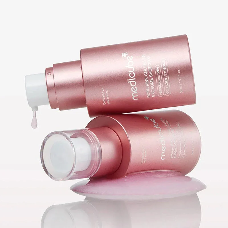 Two metallic pink bottles of Medicube PDRN Pink Collagen Exosome Shot 2000 resting horizontally, with one pump dispensing a light pink serum drop onto a glossy liquid surface.